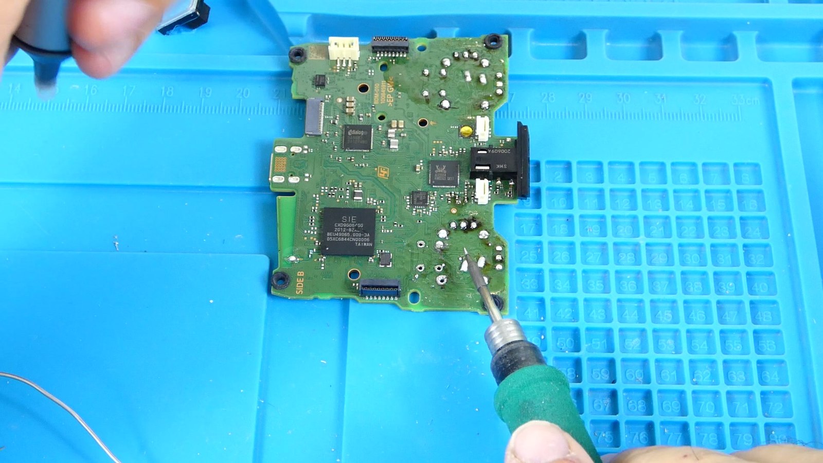 Vista cenital de la placa base de un mando DualSense mostrando chips SMD de distintos tamaños, un conector y pines densos, con el soldador aproximándose