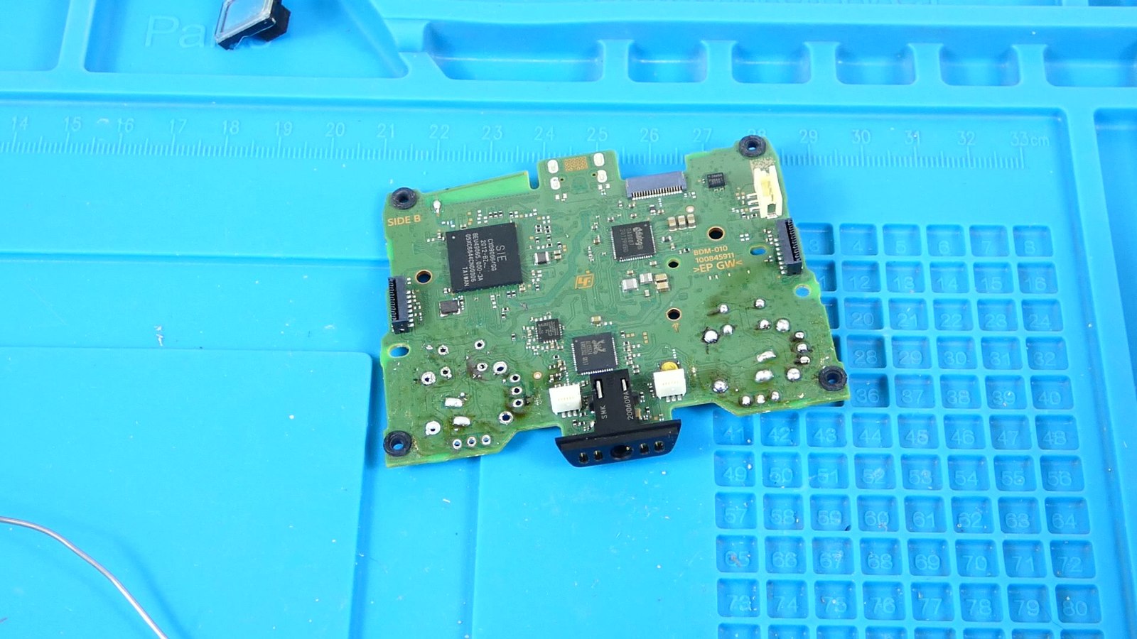 Placa base de un mando DualSense completamente desoldada y retirada, mostrando conectores, chips y pads limpios sobre una alfombrilla de trabajo con regla