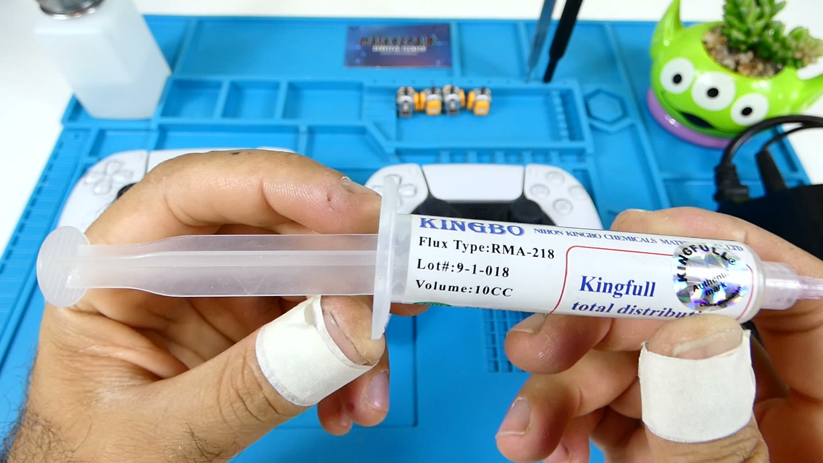 Jeringuilla de flux en gel Kingbo RMA-218 de 10cc sostenida en primer plano junto a un mando DualSense