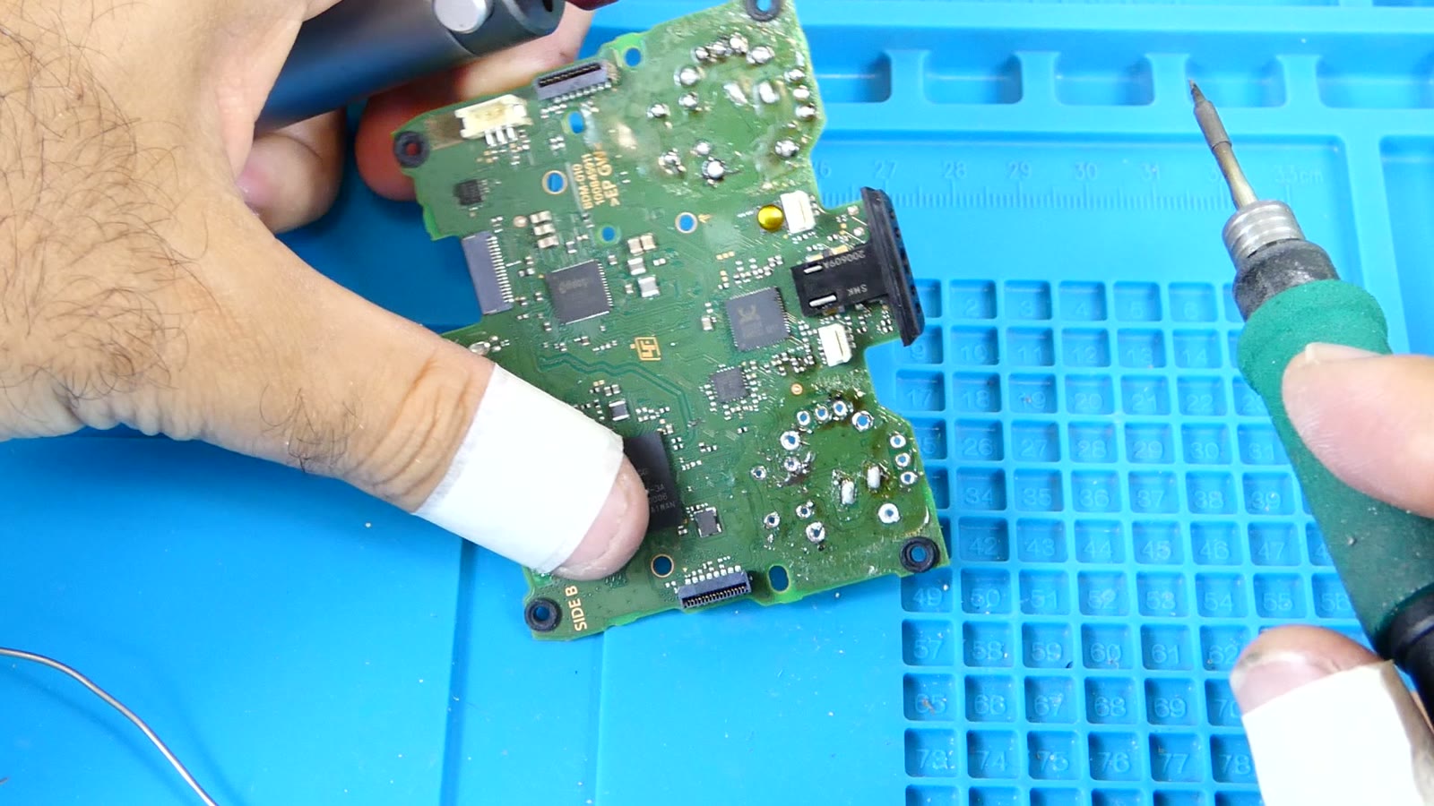 Vista cenital de una mano soldando con precisión una placa base de PS4, con el soldador de punta fina apoyado sobre un pad junto a un conector, dedo protegido con cinta